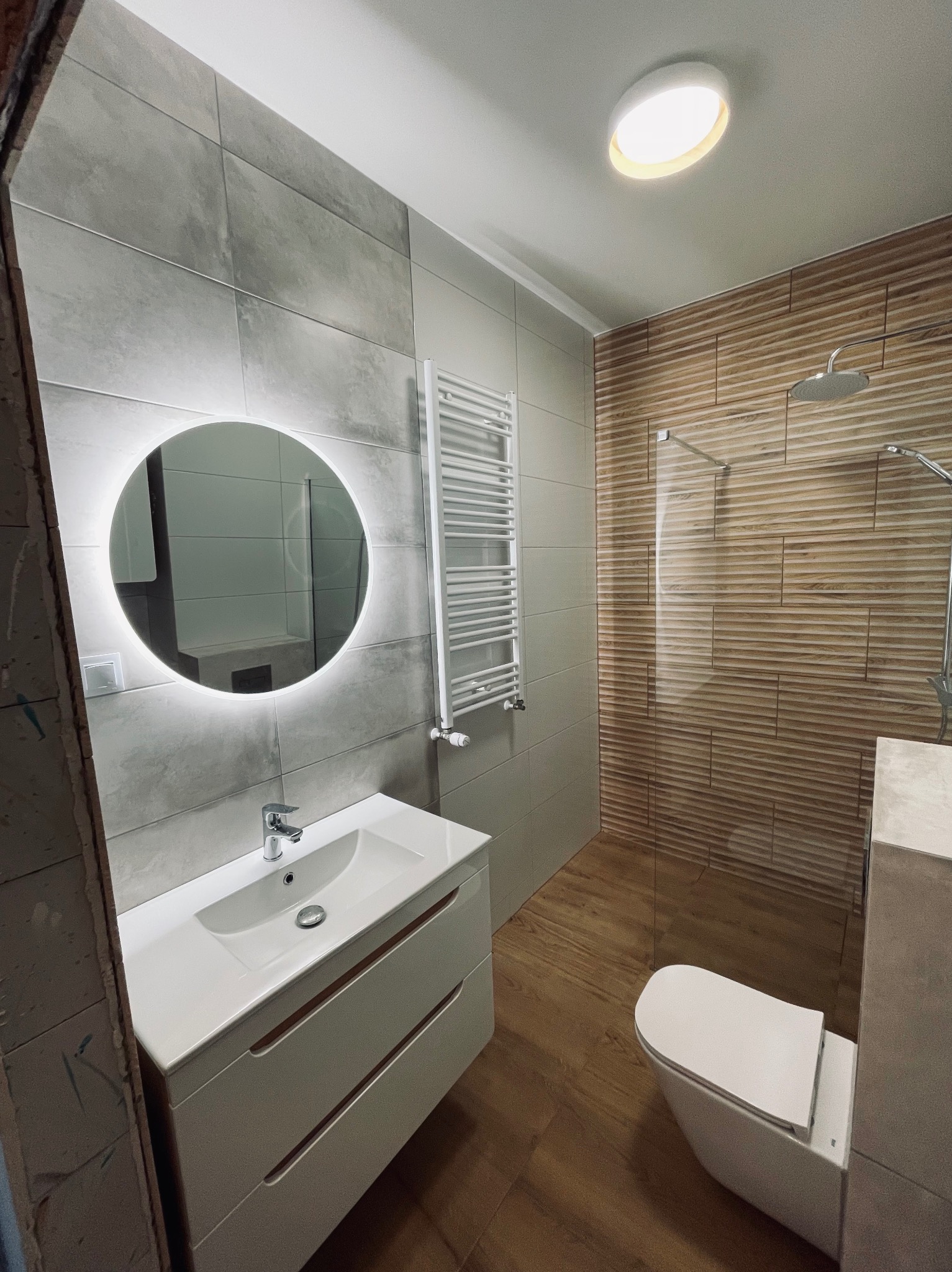 Nowoczesna łazienka po remoncie: szare płytki, okrągłe lustro z podświetleniem LED, drewnopodobne płytki pod prysznicem, biała umywalka z szafką i grzejnik drabinkowy.