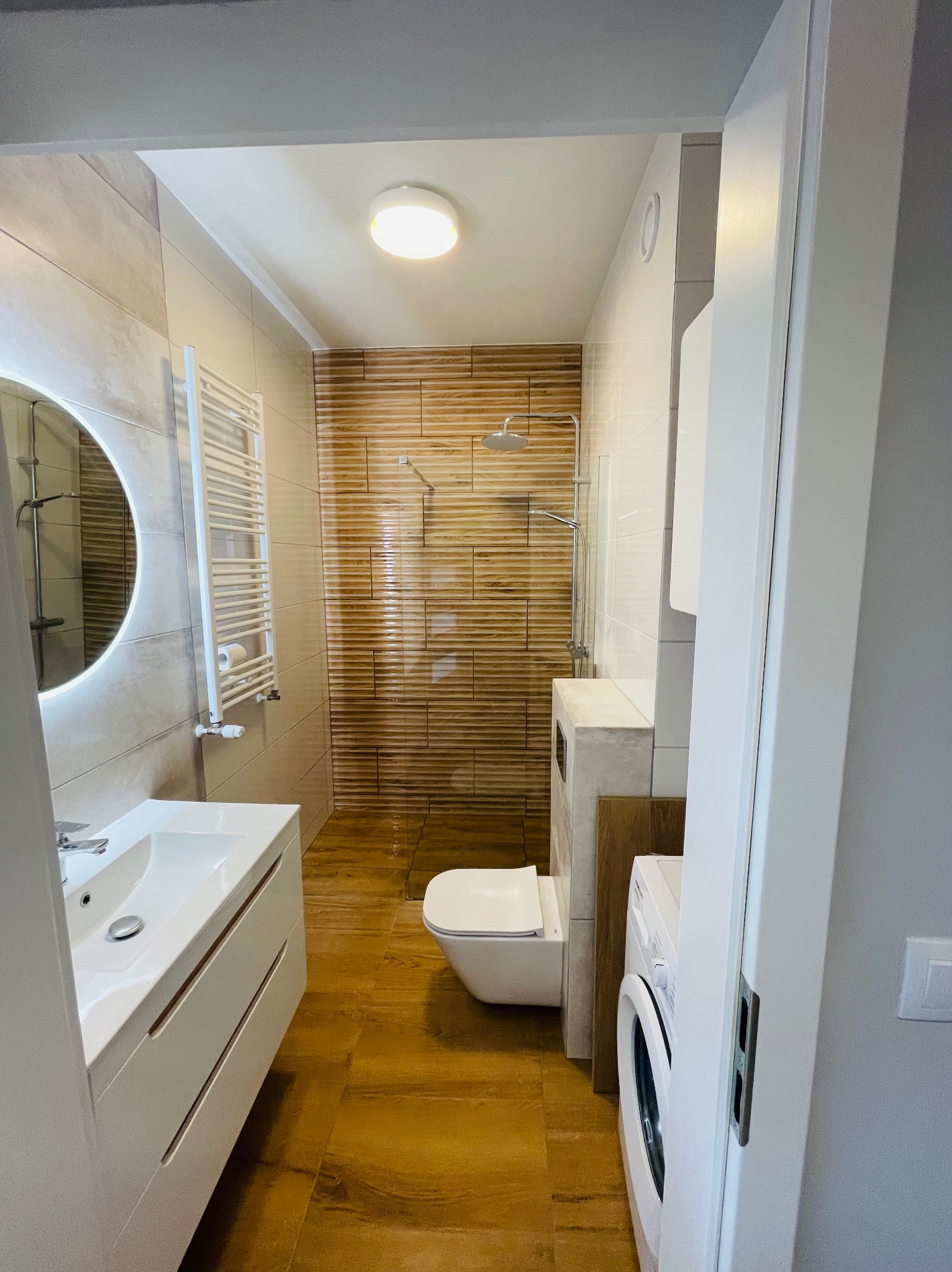 Nowoczesna łazienka z kabiną prysznicową wyłożoną płytkami imitującymi drewno, okrągłe lustro z podświetleniem i biała umywalka z szafką. Widok zza futryny.