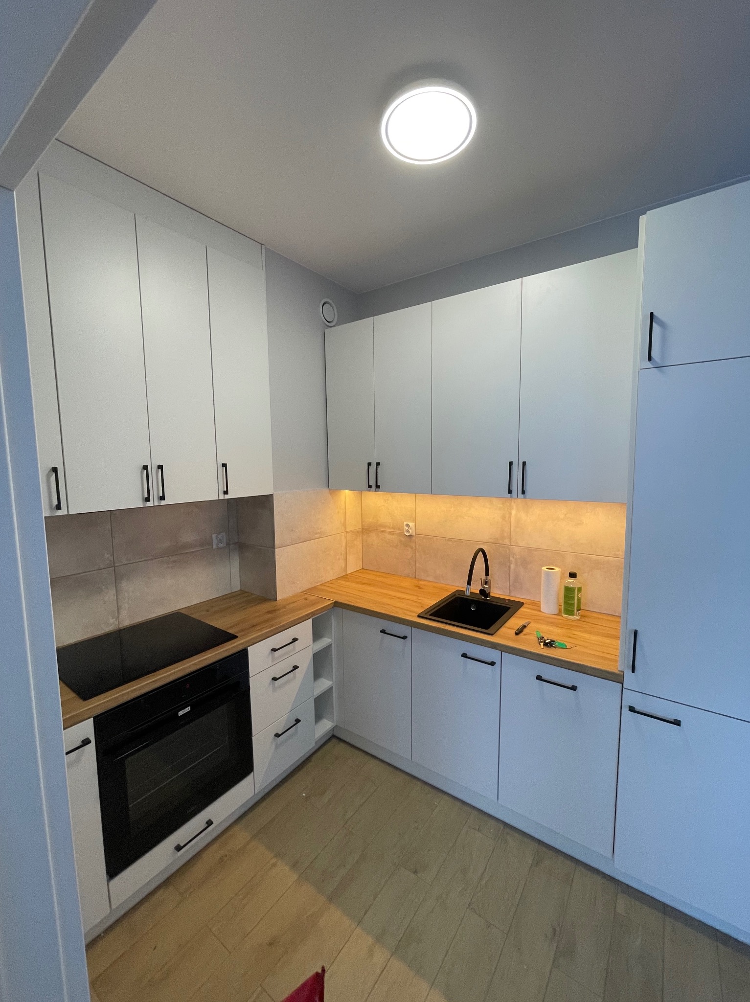 Jasna kuchnia z drewnianym blatem, czarnym zlewem i płytą indukcyjną. Nad blatem beżowe płytki. Minimalistyczne szafki z podświetleniem LED. Nowoczesne wykończenie.