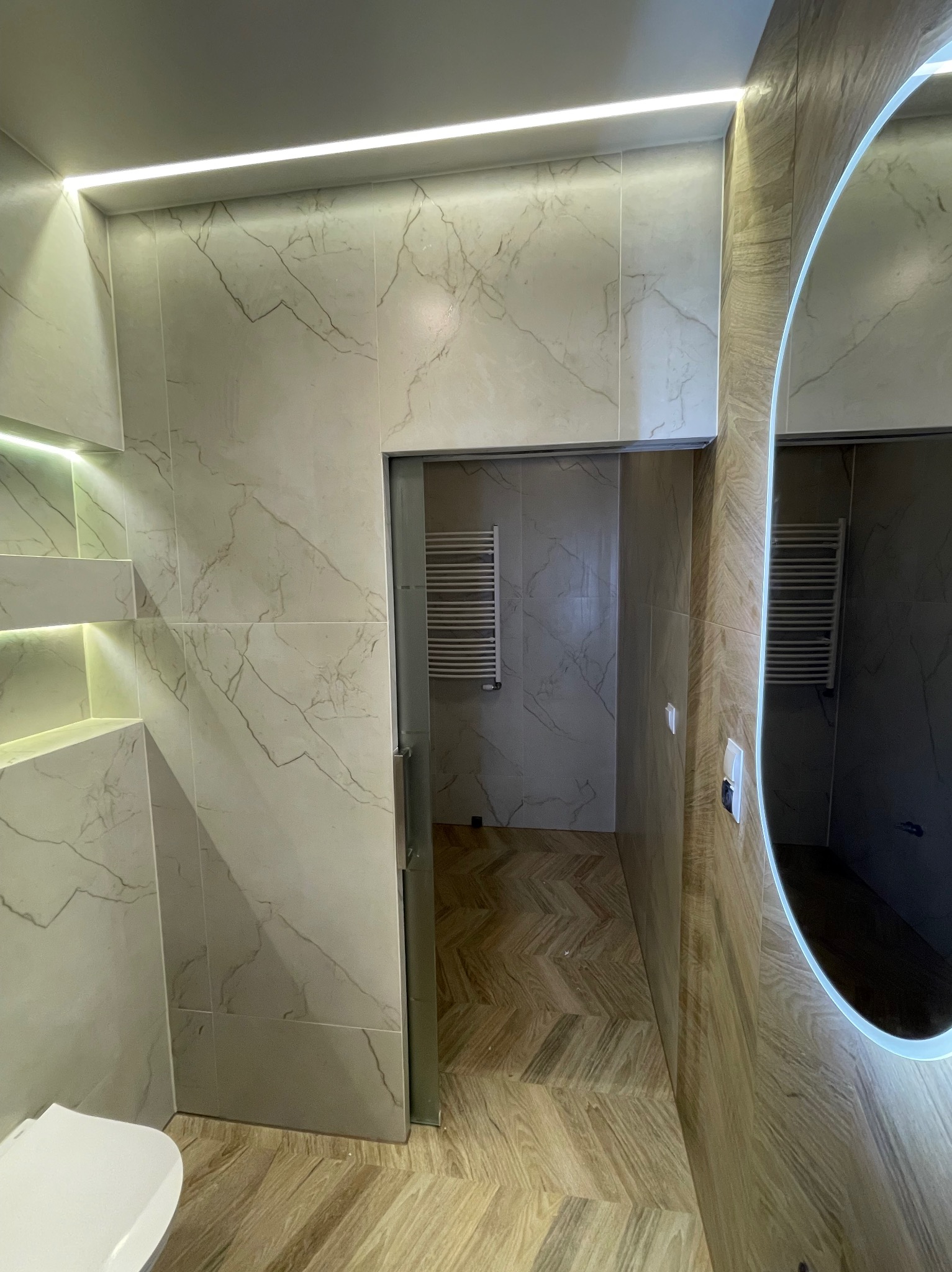 Elegancka łazienka z marmurowymi płytkami, oświetleniem LED i lustrem z podświetleniem. Widoczny fragment toalety i wzór parkietu w jodełkę na podłodze.