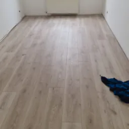 Jasne panele podłogowe ułożone w pustym pokoju z białymi ścianami i grzejnikiem pod oknem, na podłodze leży niebieska bluza.