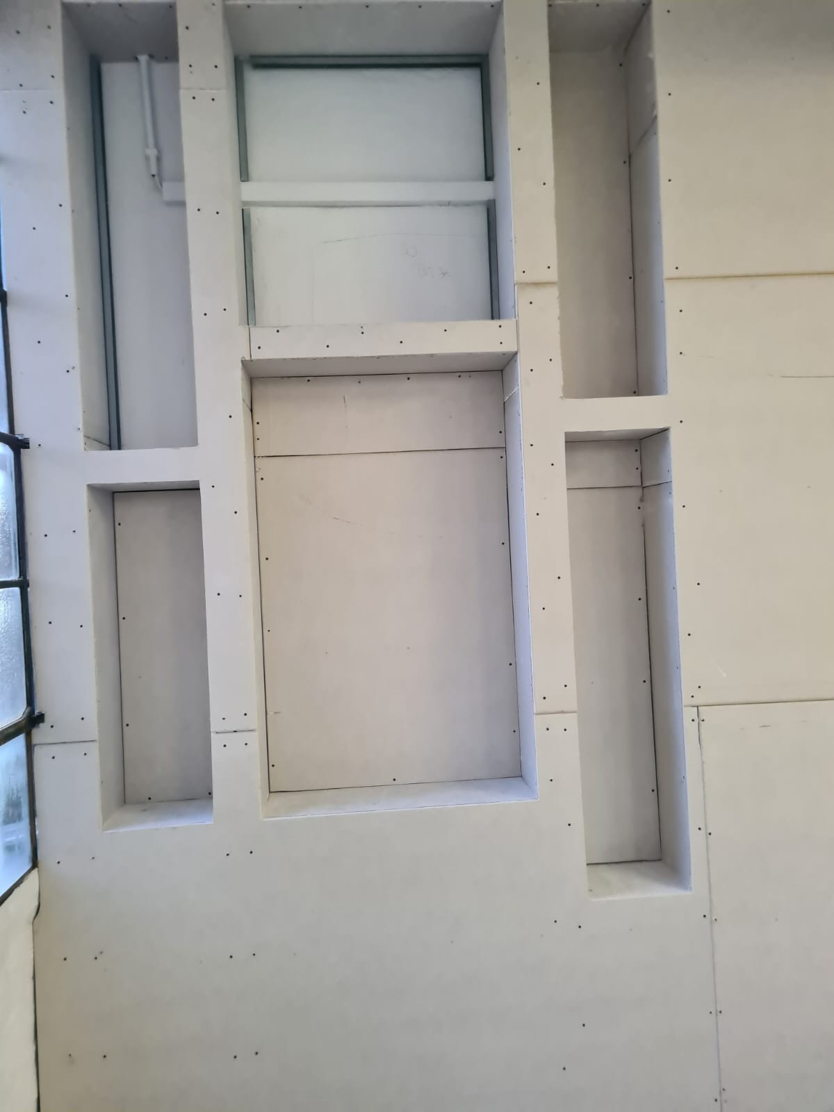 Ściana z zamontowanymi płytami gipsowo-kartonowymi, tworzącymi wnęki o różnych kształtach i rozmiarach, widoczne ślady po wkrętach, surowy stan przed wykończeniem.