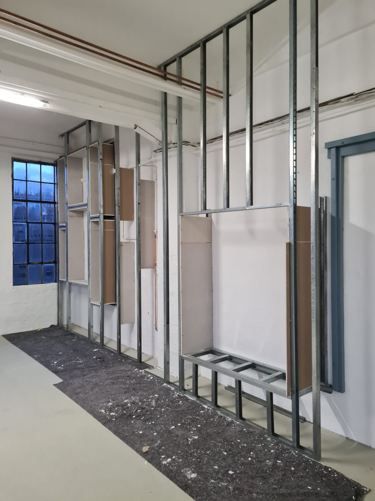 Metalowa konstrukcja szkieletowa z fragmentami płyt kartonowo-gipsowych, tworząca zabudowę wnęki w pomieszczeniu z dużym oknem i zabezpieczoną podłogą.