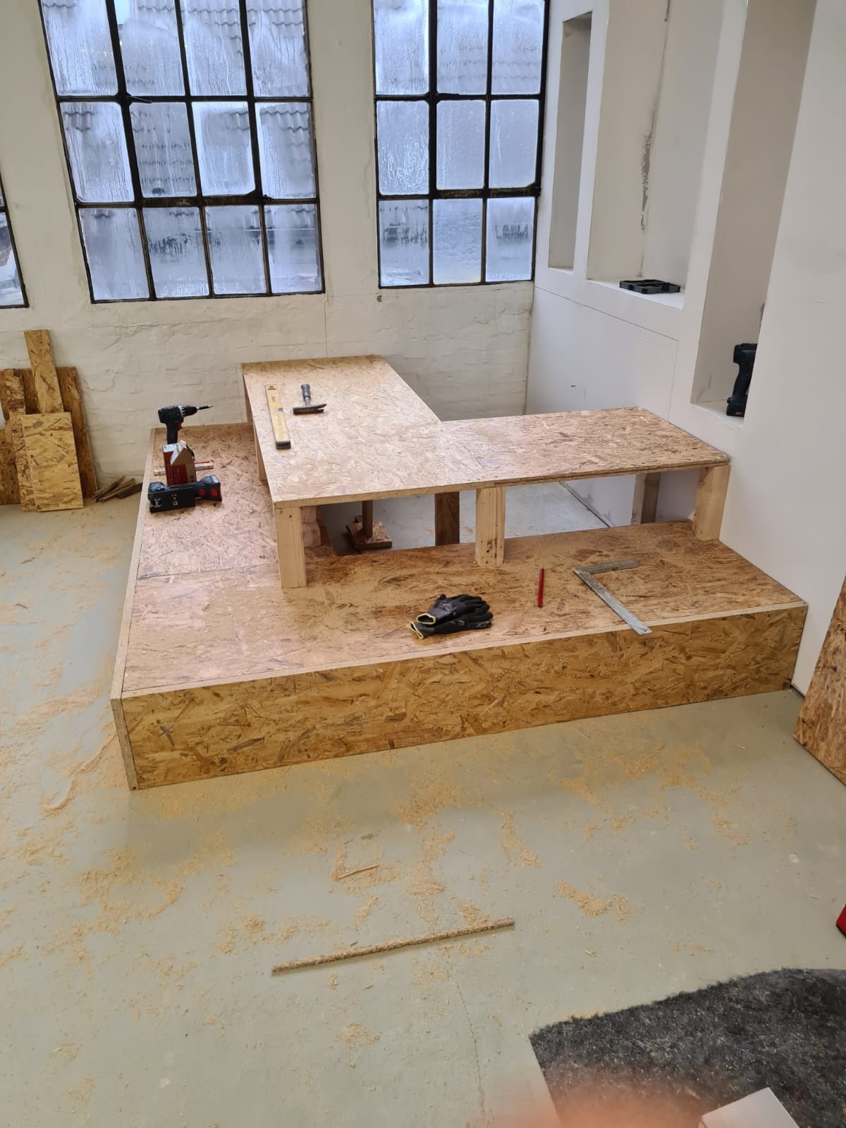 Wnętrze w trakcie zabudowy z płyty OSB: konstrukcja w kształcie litery L, narzędzia (wiertarka, poziomica, kątownik), rękawice robocze, widoczne wióry na podłodze, duże okna w tle.