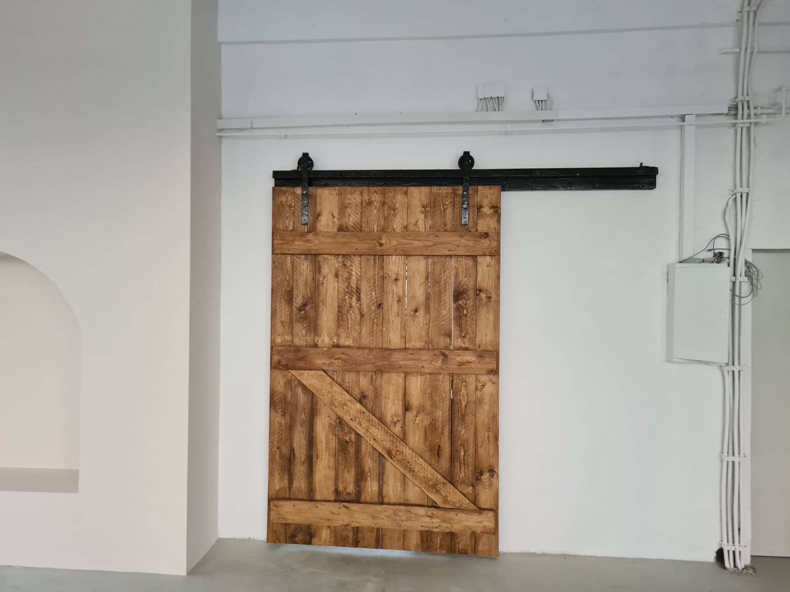 Drewniane drzwi przesuwne w stylu rustykalnym na czarnym metalowym prowadzeniu, zamontowane w białym wnętrzu z widocznymi przewodami elektrycznymi.