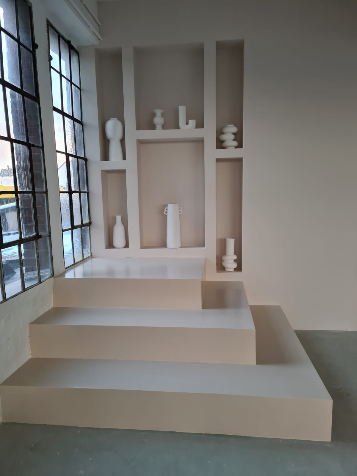 Minimalistyczne wnętrze z beżowymi ścianami, wnękami z dekoracyjnymi wazonami i schodkowym podestem, oświetlone przez duże okno z czarnymi szprosami.