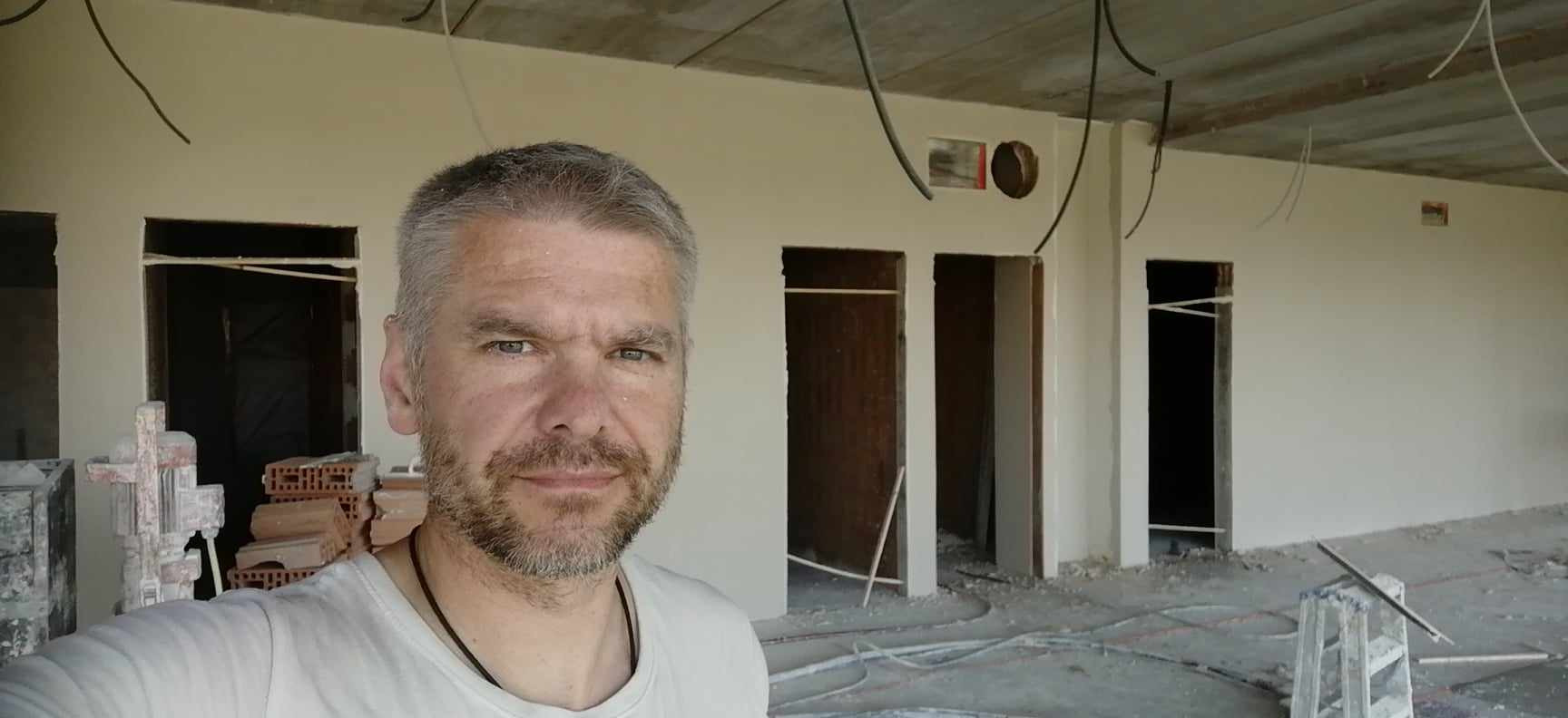 Mężczyzna z siwiejącą brodą pozuje na tle świeżo otynkowanych ścian w surowym stanie, widoczne instalacje elektryczne na suficie i resztki materiałów budowlanych na podłodze.