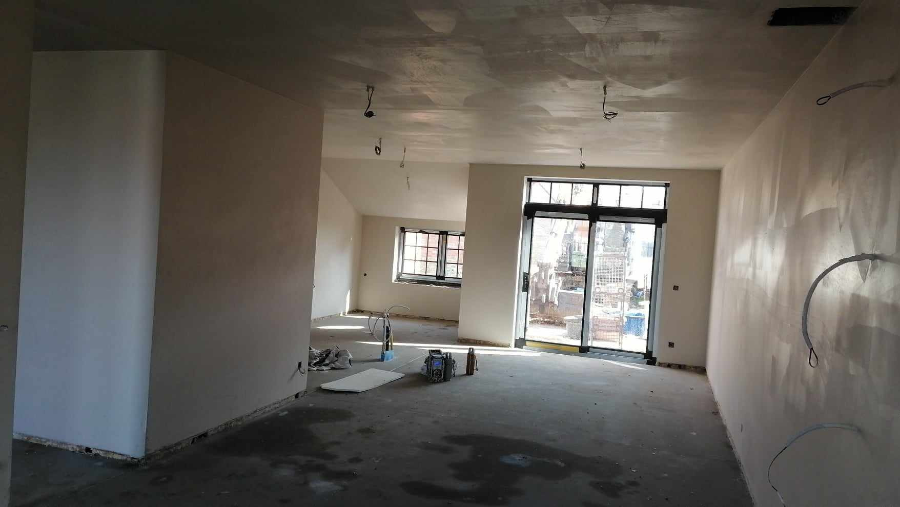 Wnętrze domu w trakcie prac wykończeniowych: świeżo nałożone, surowe tynki na ścianach i suficie, widoczne kable elektryczne, sprzęt tynkarski, okna z widokiem na zewnątrz.