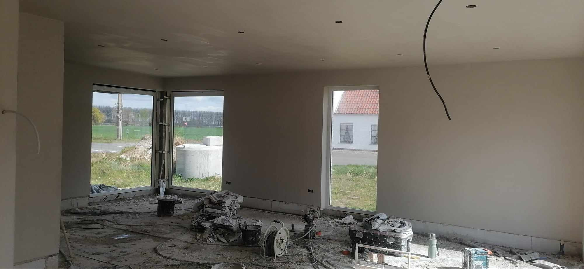 Wnętrze w trakcie prac wykończeniowych: świeżo otynkowane ściany, podłoga zabezpieczona folią, widoczne worki z zaprawą, narzędzia i przewody, duże okna z widokiem na zewnątrz.