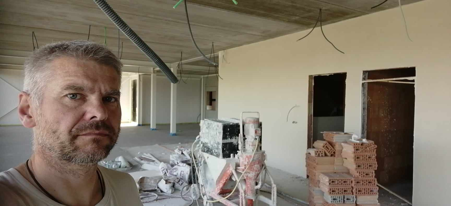 Mężczyzna robiący selfie na tle pomieszczenia w trakcie prac tynkarskich, widoczna maszyna do tynkowania i sterta cegieł