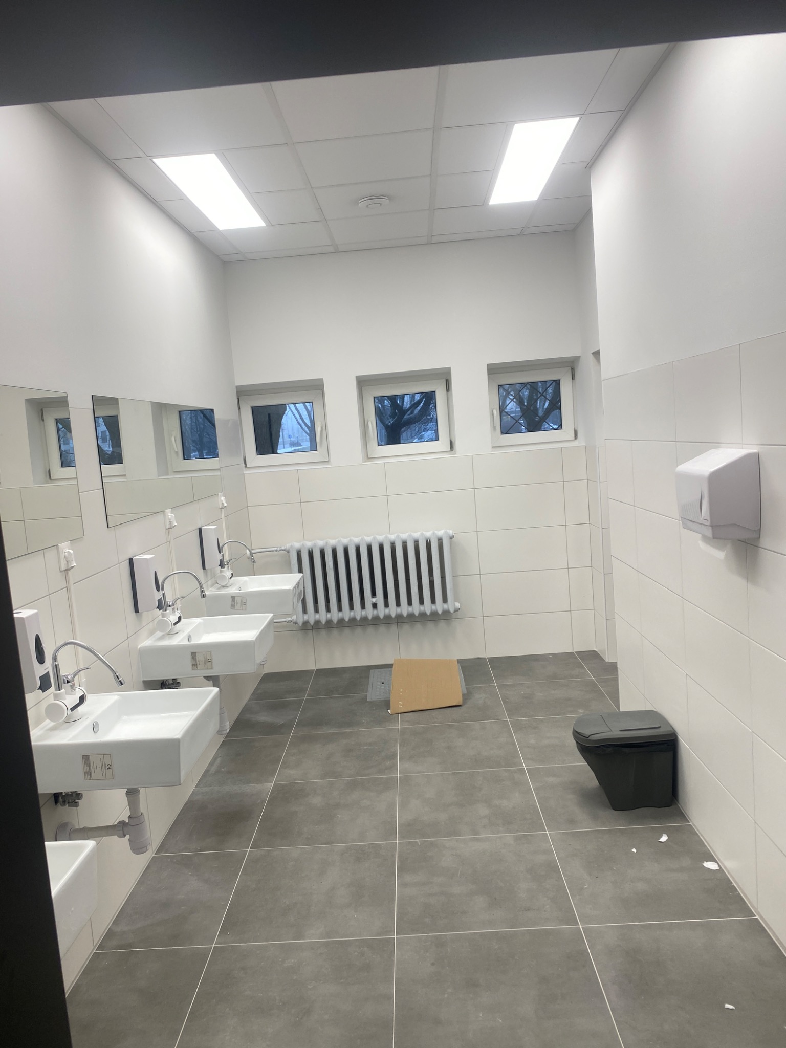 Wnętrze publicznej toalety po remoncie: rzędy białych umywalek z chromowanymi kranami, białe płytki na ścianach, szare płytki na podłodze, grzejnik żeberkowy pod oknami, karton na podłodze, kosz...
