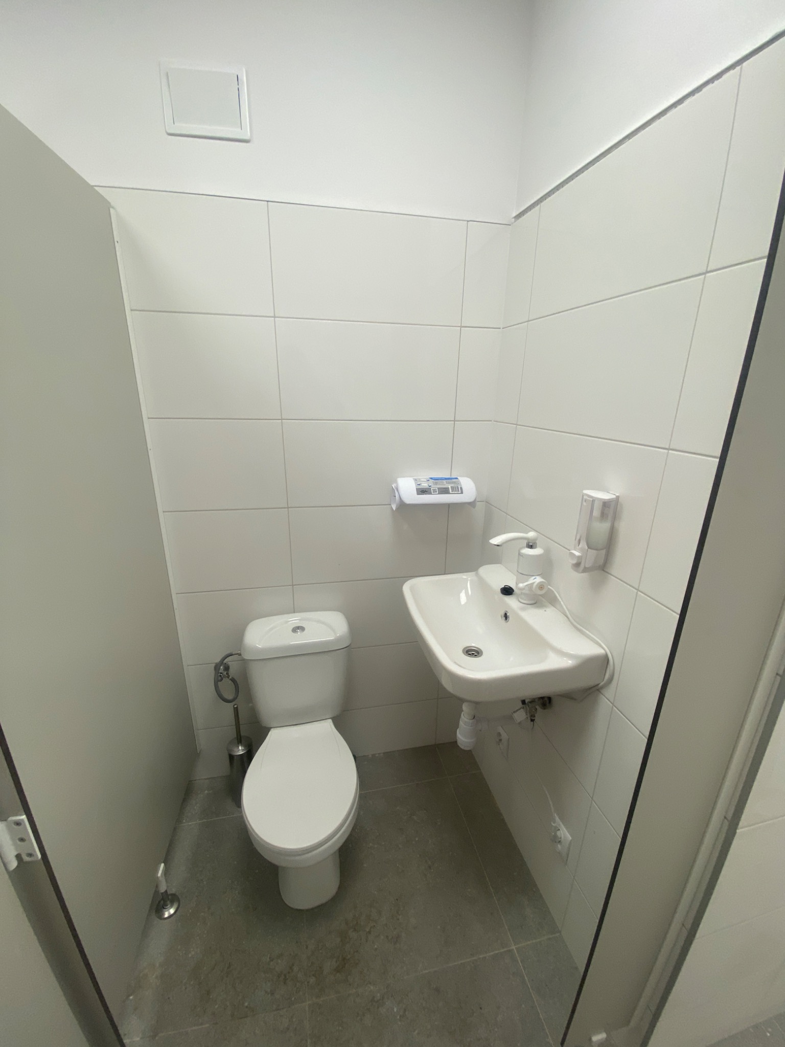 Wnętrze małej, jasnej łazienki z białą ceramiką, płytkami i drzwiami, widok z perspektywy wejścia, z widoczną toaletą, umywalką z dozownikiem mydła i uchwytem na papier toaletowy.