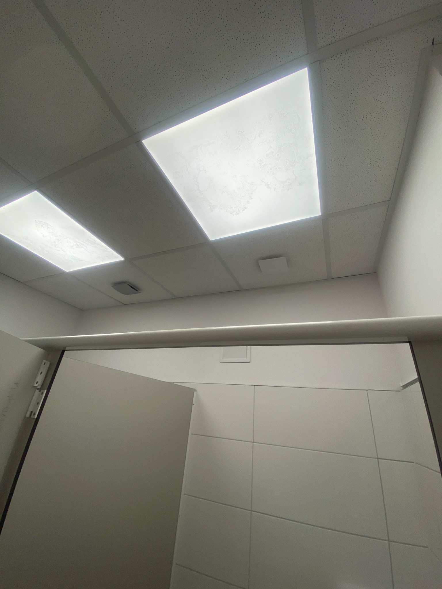 Wykończona łazienka w biurze z białymi płytkami na ścianach, widok na sufit z podwieszanymi panelami i oświetleniem LED.