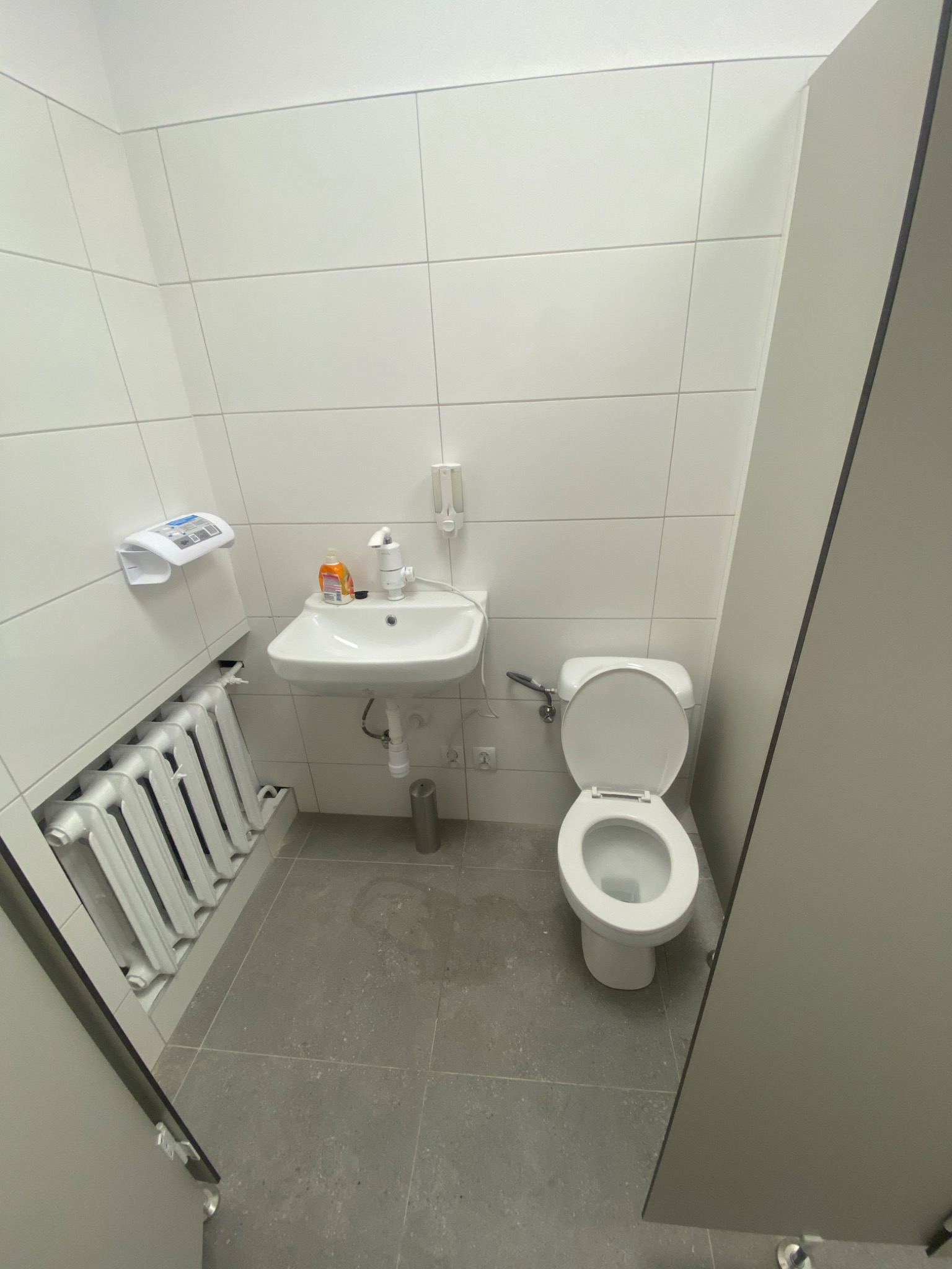 Wnętrze łazienki z białą umywalką, toaletą z otwartą klapą, grzejnikiem żeberkowym i szarą podłogą z płytek, widziane z perspektywy drzwi kabiny.