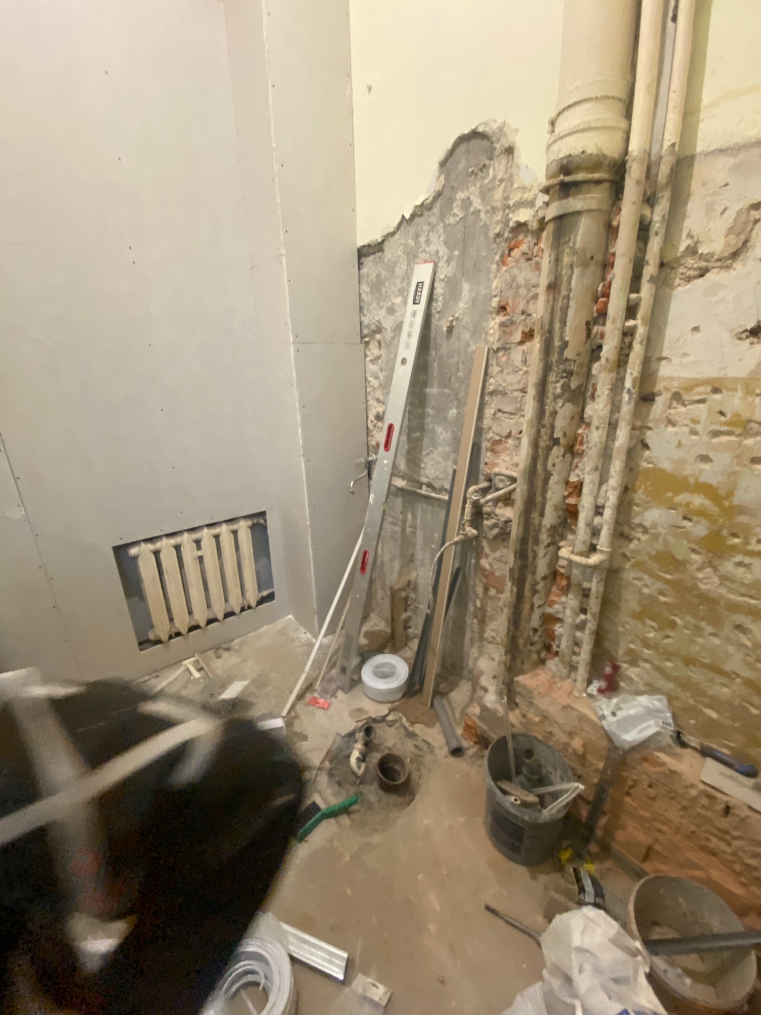 Łazienka w trakcie remontu: ściana z płyt kartonowo-gipsowych z widocznym grzejnikiem, fragment odsłoniętej ściany z cegły i rurami, poziomica oparta o ścianę, widoczne narzędzia i wiaderka.