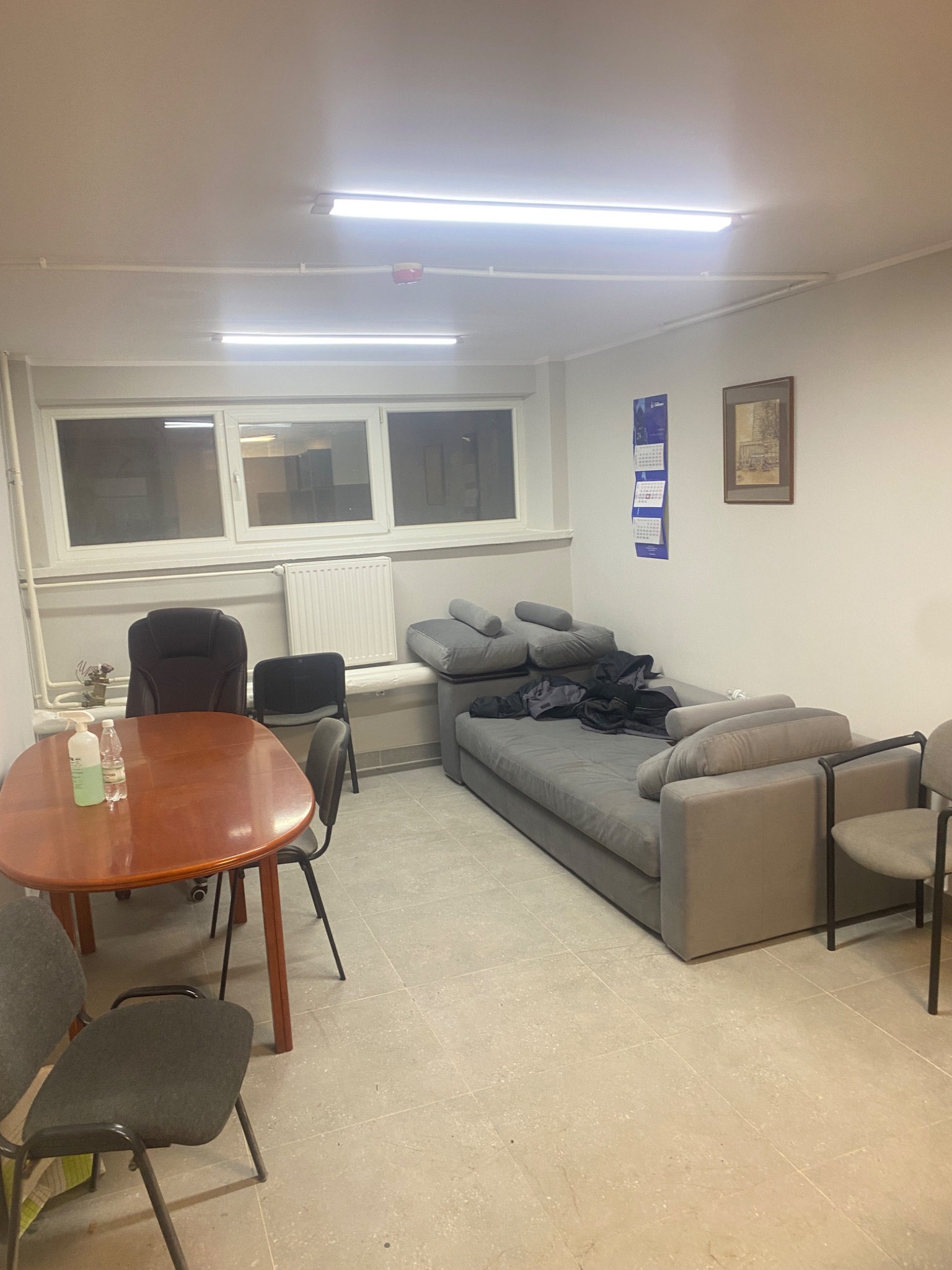 Zdjęcie przedstawia wnętrze biura z drewnianym stołem, krzesłami, szarą sofą i kalendarzem na ścianie, ukazujące potencjalne miejsce pracy po zakończeniu prac wykończeniowych.