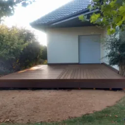 Nowoczesny taras z desek w kolorze ciemnego drewna, przylegający do budynku z szarą roletą okienną, otoczony piaskiem i zielenią, z widocznymi narzędziami ogrodniczymi w rogu.