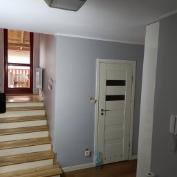 Usługi i Inwestycje Mateusz Janicki - Korytarz w trakcie odświeżania: pomalowane na szaro ściany, białe drzwi z poziomymi przeszkleniami, drewniane schody prowadzące na zewnątrz, butelka wody mineralnej przy ścianie.