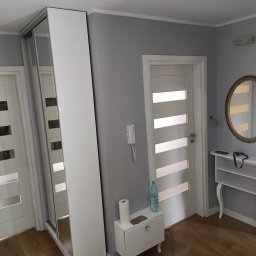 Usługi i Inwestycje Mateusz Janicki - Jasne wnętrze przedpokoju z przesuwną szafą z lustrem, białymi drzwiami z poziomymi przeszkleniami, owalnym lustrem w złotej ramie i białą konsolką pod lustrem, na podłodze panele w kolorze drewna.