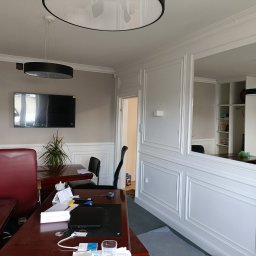 Usługi i Inwestycje Mateusz Janicki - Wykończone biuro z białą boazerią panelową, lustrem w ścianie, ciemnymi biurkami i czerwonym krzesłem, widoczny telewizor na ścianie i lampy sufitowe.