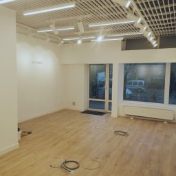 Usługi i Inwestycje Mateusz Janicki - Jasne, przestronne wnętrze biurowe przed wykończeniem, z drewnianą podłogą, białymi ścianami i nowoczesnym oświetleniem sufitowym, widoczne przewody elektryczne na podłodze.