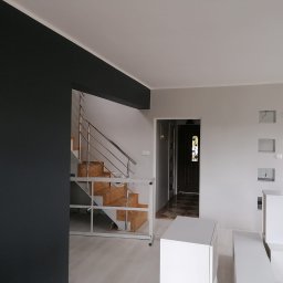 Usługi i Inwestycje Mateusz Janicki - Wnętrze domu po remoncie, z pomalowaną na grafitowo ścianą, drewnianymi schodami z metalową balustradą, białymi ścianami i elementami dekoracyjnymi z płyt kartonowo-gipsowych oraz widocznymi...