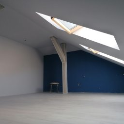 Usługi i Inwestycje Mateusz Janicki - Poddasze po odświeżeniu: białe ściany kontrastują z granatową ścianą akcentową, drewniane okna dachowe wpuszczają światło, a proste krzesło dodaje minimalistycznego charakteru.