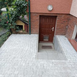 Usługi i Inwestycje Mateusz Janicki - Ułożona kostka brukowa prowadząca do wejścia do piwnicy w domu z elewacją z cegły klinkierowej, widoczny fragment okna piwnicznego i buda dla psa w tle.