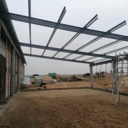 Usługi i Inwestycje Mateusz Janicki - Konstrukcja stalowa z profili zamkniętych tworząca zadaszenie w trakcie budowy, widoczne kolumny betonowe i teren budowy.