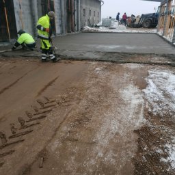 Usługi i Inwestycje Mateusz Janicki - Wyrównywanie wylewki betonowej w hali produkcyjnej; dwóch robotników w odblaskowych ubraniach, jeden klęczy z łatą, drugi wyrównuje betonową powierzchnię, w tle widoczny traktor i materiały budowlane.