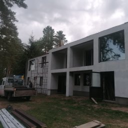 Usługi i Inwestycje Mateusz Janicki - Surowy, dwupiętrowy dom z oknami w trakcie wykończenia elewacji zewnętrznej, widoczny częściowo zakryty rusztowaniem, z transportowym samochodem dostawczym na posesji i elementami budowlanymi...