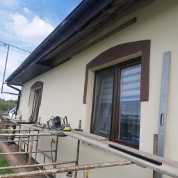 Usługi i Inwestycje Mateusz Janicki - Elewacja domu z beżowym tynkiem, brązowymi obramowaniami okien i rusztowaniem, na którym leżą narzędzia i poziomica.