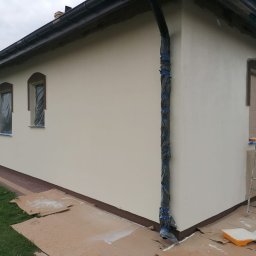 Usługi i Inwestycje Mateusz Janicki - Elewacja domu jednorodzinnego w trakcie malowania, okna zabezpieczone folią, rynna owinięta czarną folią i taśmą, widoczne kartony ochronne na trawie, drabina oparta o ścianę.