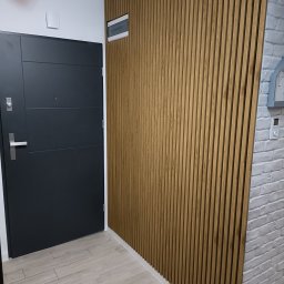 Usługi i Inwestycje Mateusz Janicki - Nowoczesny przedpokój z drewnianą ścianą z lameli, szara drzwi wejściowe i podłoga imitująca drewno, obok ściana z cegły pomalowana na biało.