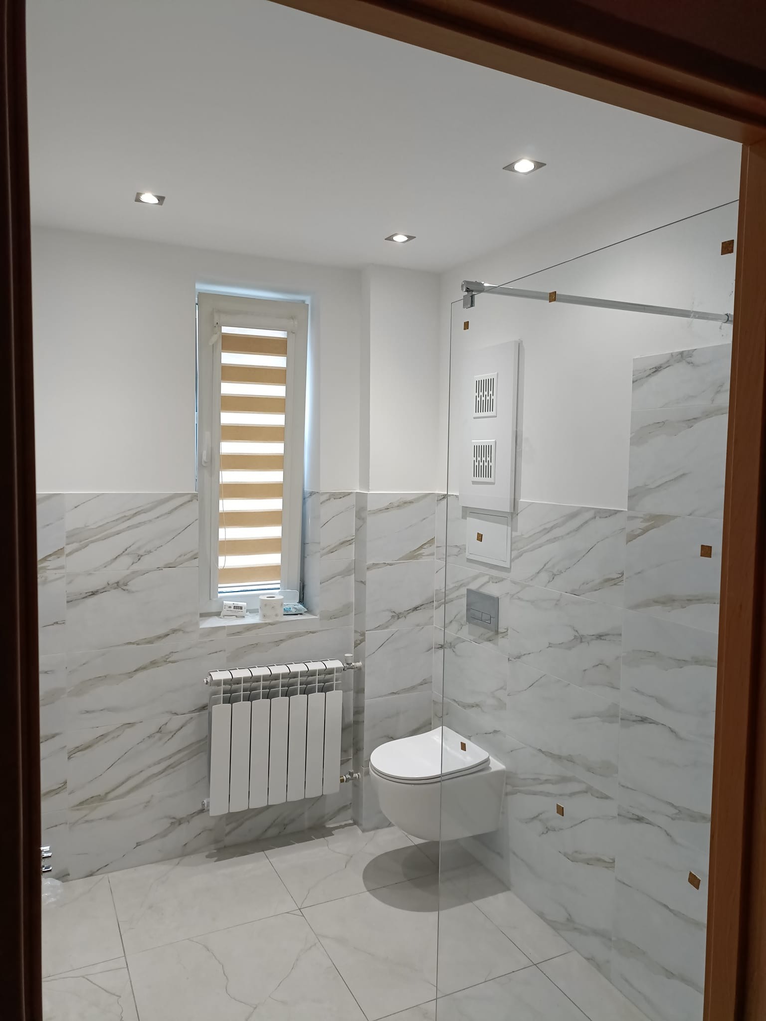 Nowoczesna łazienka z marmurowymi płytkami, podwieszaną toaletą, grzejnikiem panelowym i kabiną prysznicową ze szklaną ścianką.