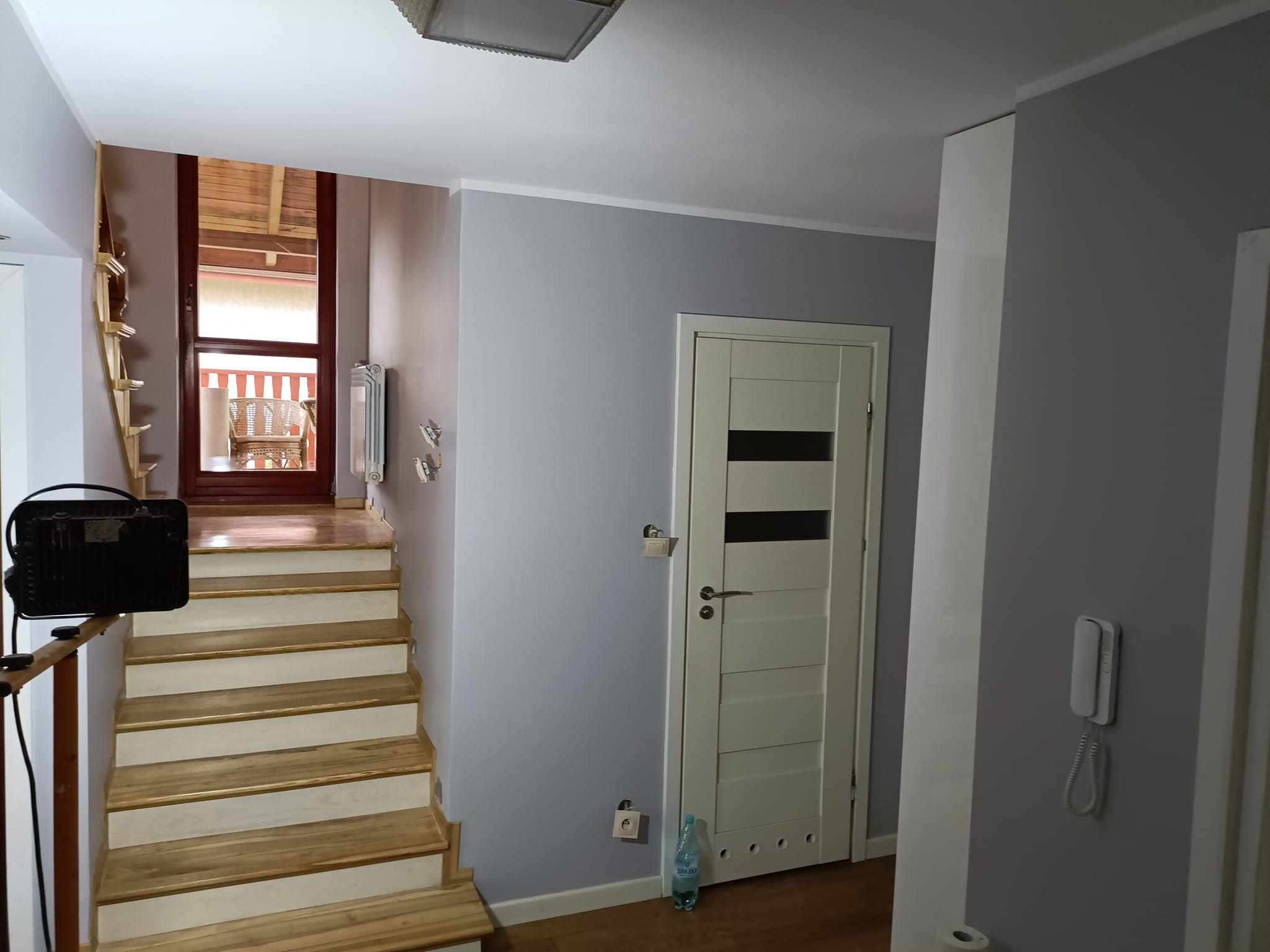 Korytarz w trakcie odświeżania: pomalowane na szaro ściany, białe drzwi z poziomymi przeszkleniami, drewniane schody prowadzące na zewnątrz, butelka wody mineralnej przy ścianie.