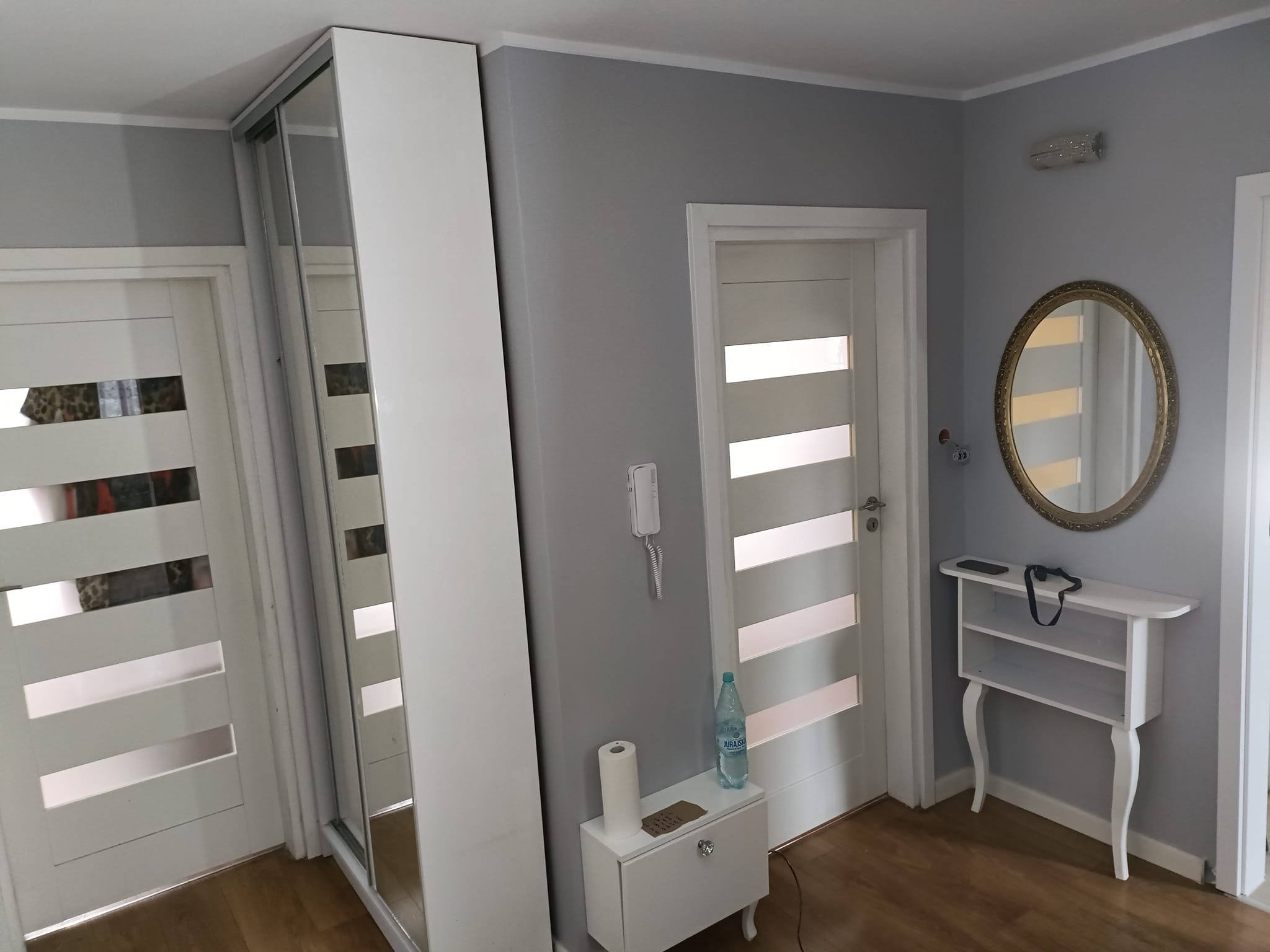 Jasne wnętrze przedpokoju z przesuwną szafą z lustrem, białymi drzwiami z poziomymi przeszkleniami, owalnym lustrem w złotej ramie i białą konsolką pod lustrem, na podłodze panele w kolorze drewna.