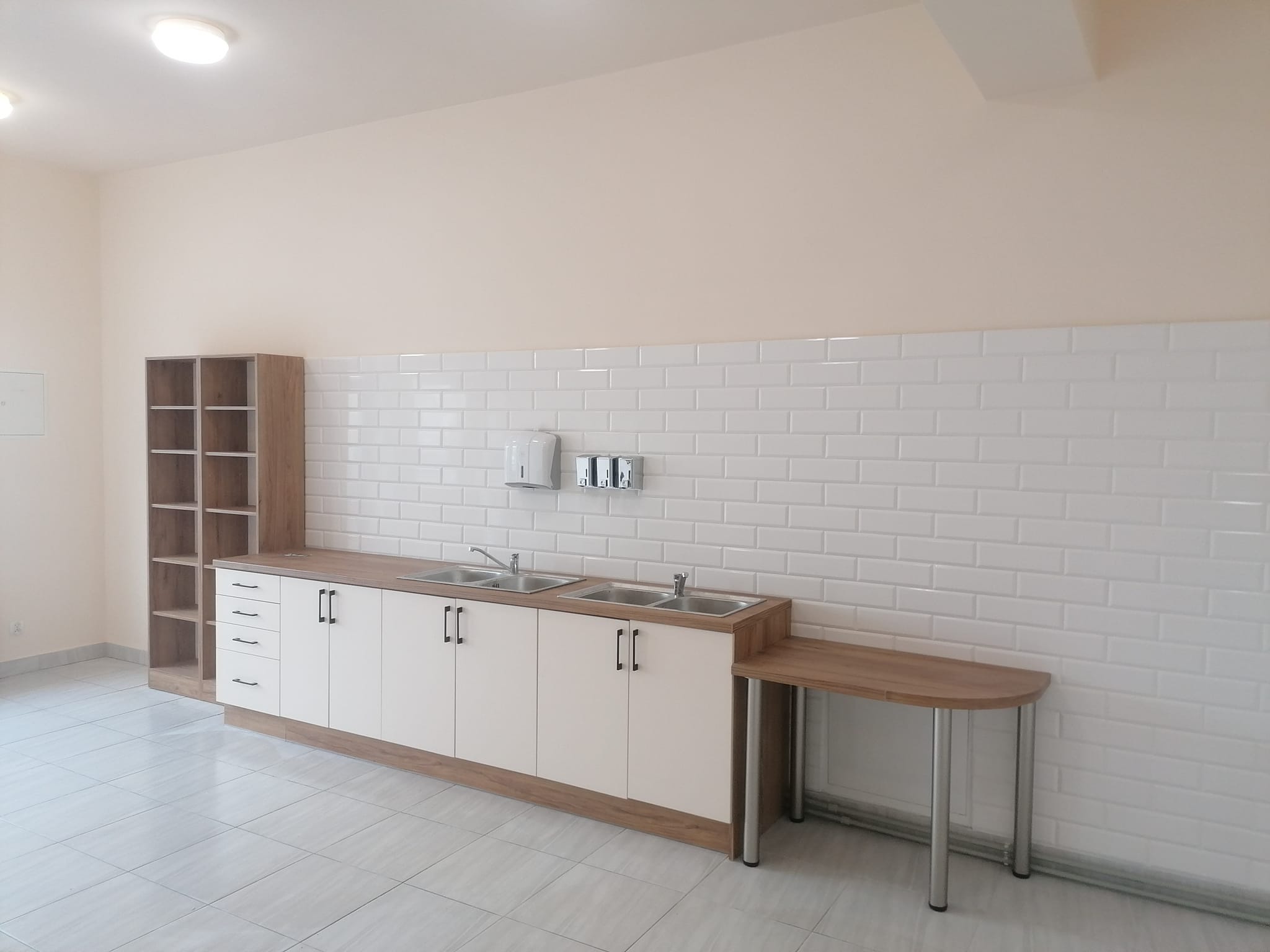 Nowoczesna, jasna umywalnia z trzema zlewozmywakami, białymi kafelkami na ścianie i podłogą w szarej tonacji, uzupełniona drewnianym regałem i półokrągłym blatem na metalowych nogach.