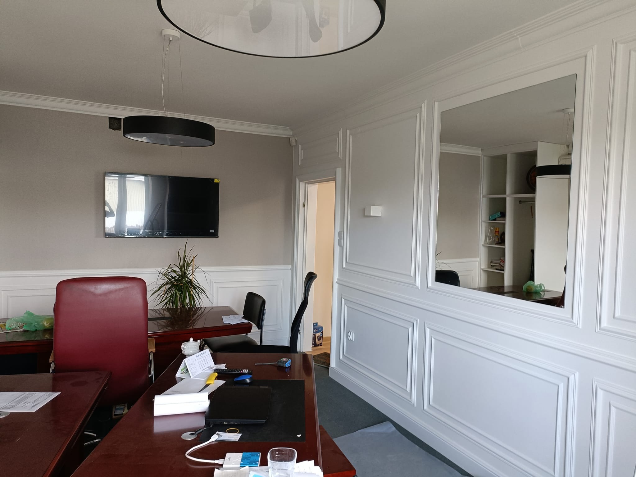 Wykończone biuro z białą boazerią panelową, lustrem w ścianie, ciemnymi biurkami i czerwonym krzesłem, widoczny telewizor na ścianie i lampy sufitowe.