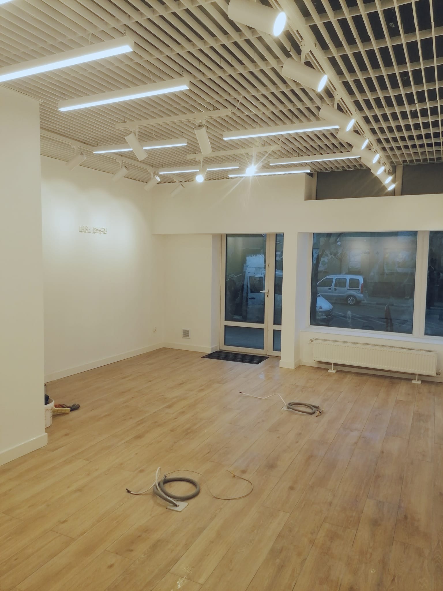 Jasne, przestronne wnętrze biurowe przed wykończeniem, z drewnianą podłogą, białymi ścianami i nowoczesnym oświetleniem sufitowym, widoczne przewody elektryczne na podłodze.