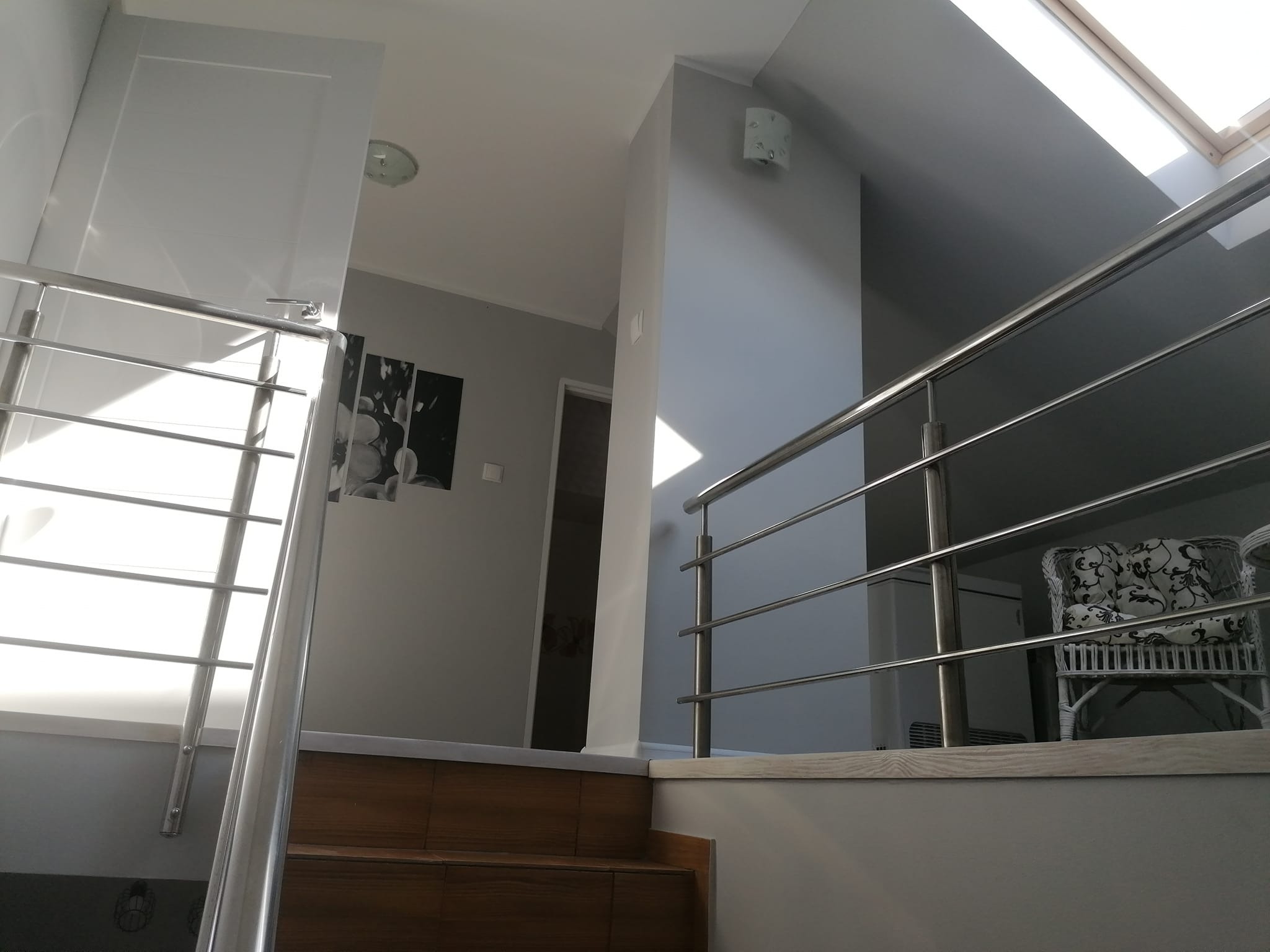 Wnętrze domu: schody z drewnianymi stopniami i metalową balustradą, widok na korytarz z czarno-białymi obrazami na ścianie, w tle krzesło z wzorzystą tapicerką, oświetlenie sufitowe i okno dachowe.