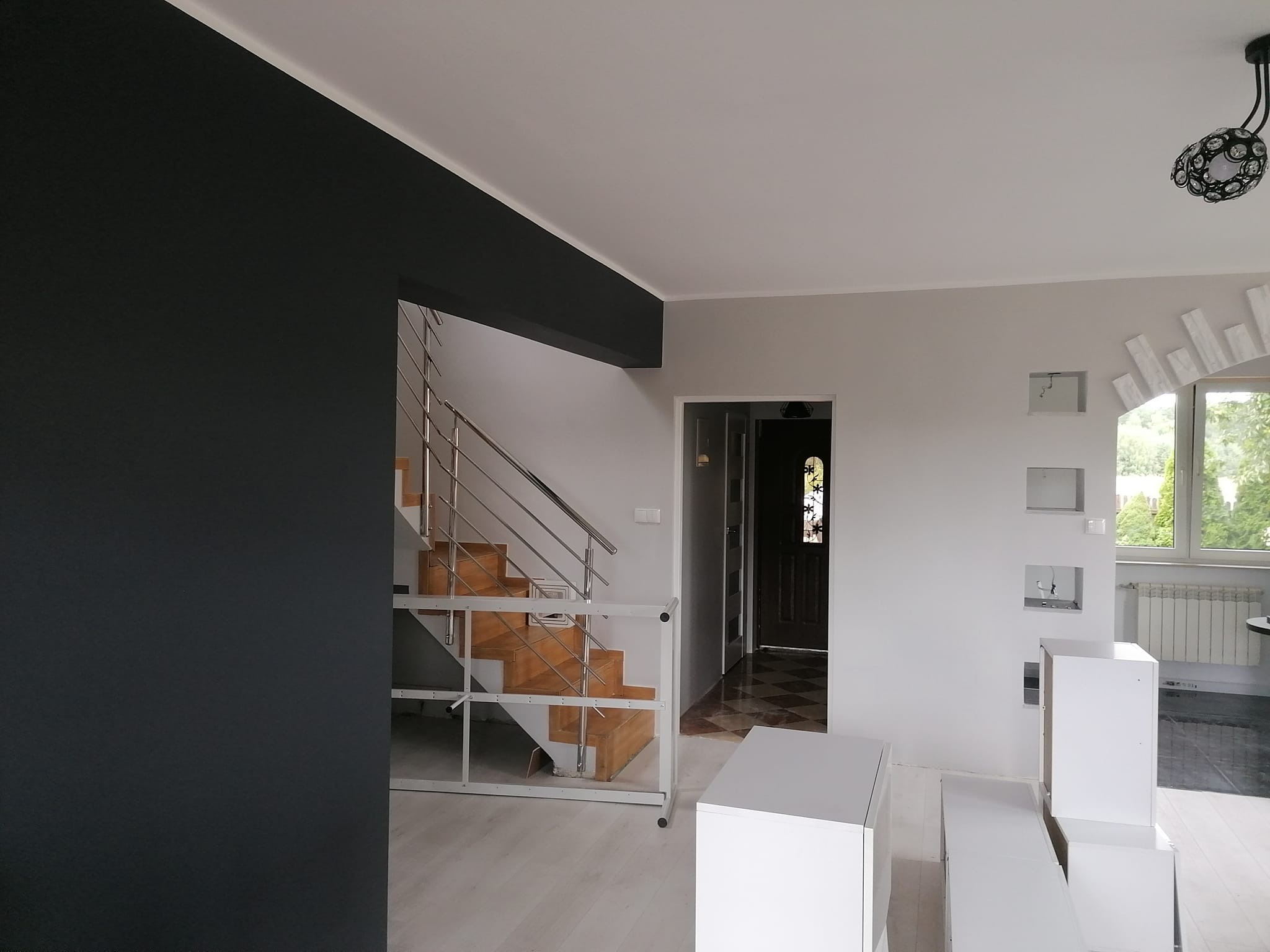 Wnętrze domu po remoncie, z pomalowaną na grafitowo ścianą, drewnianymi schodami z metalową balustradą, białymi ścianami i elementami dekoracyjnymi z płyt kartonowo-gipsowych oraz widocznymi...