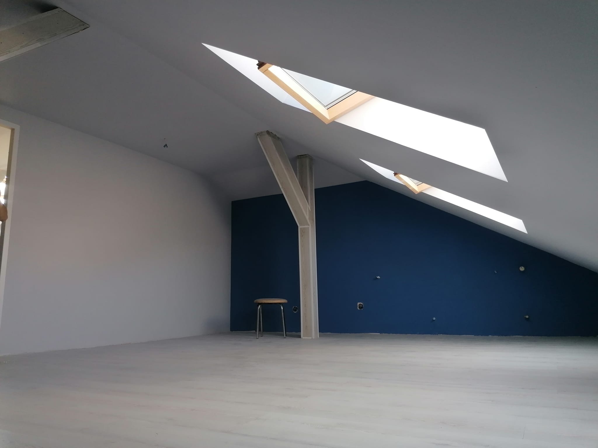 Poddasze po odświeżeniu: białe ściany kontrastują z granatową ścianą akcentową, drewniane okna dachowe wpuszczają światło, a proste krzesło dodaje minimalistycznego charakteru.