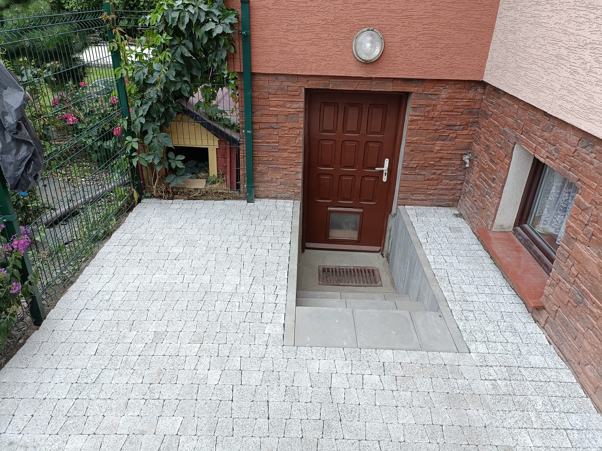 Ułożona kostka brukowa prowadząca do wejścia do piwnicy w domu z elewacją z cegły klinkierowej, widoczny fragment okna piwnicznego i buda dla psa w tle.