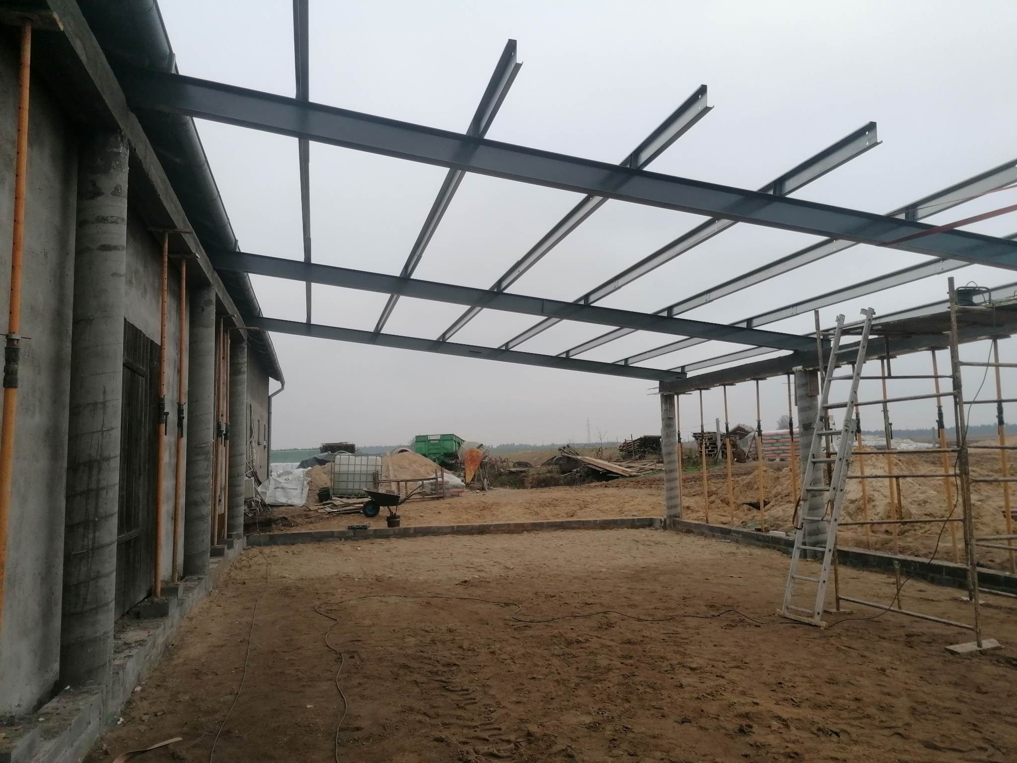 Konstrukcja stalowa z profili zamkniętych tworząca zadaszenie w trakcie budowy, widoczne kolumny betonowe i teren budowy.