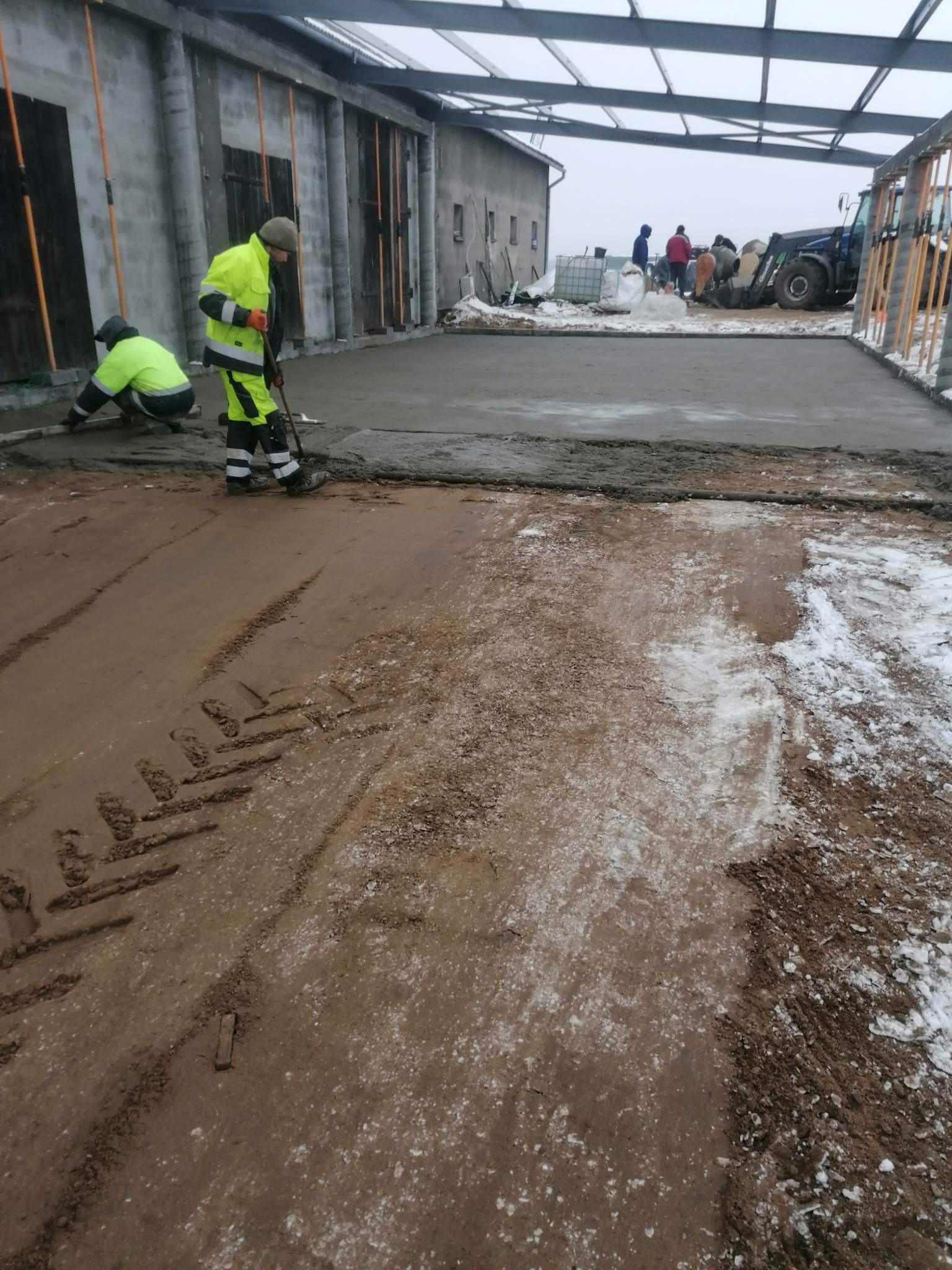 Wyrównywanie wylewki betonowej w hali produkcyjnej; dwóch robotników w odblaskowych ubraniach, jeden klęczy z łatą, drugi wyrównuje betonową powierzchnię, w tle widoczny traktor i materiały budowlane.