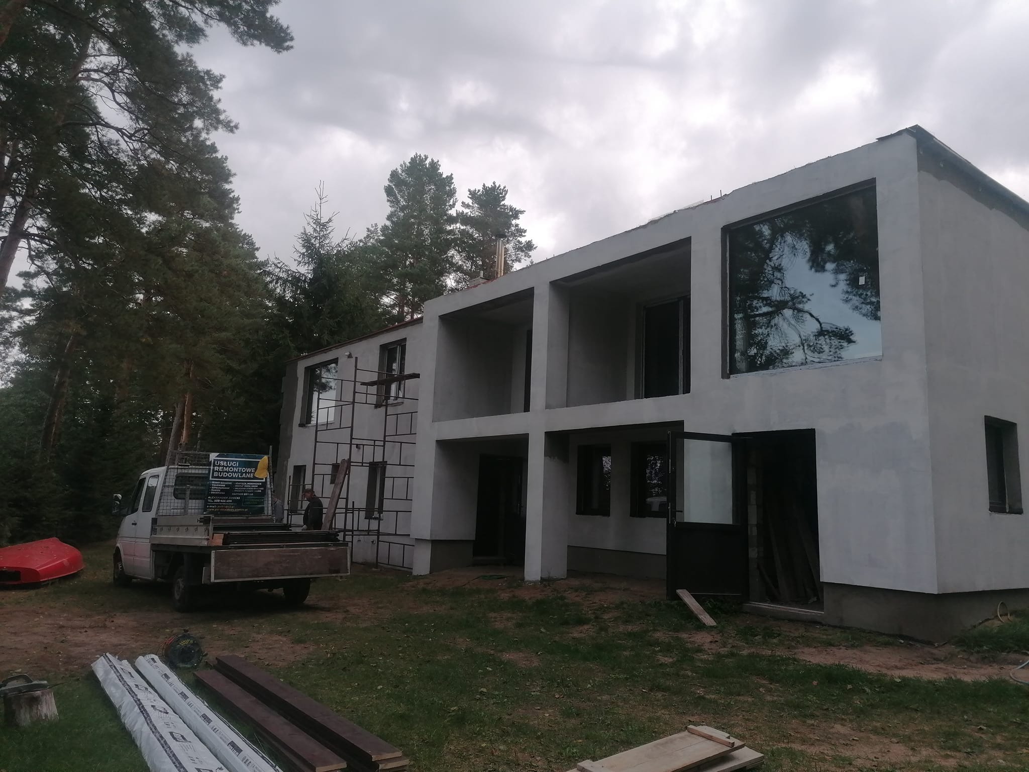 Surowy, dwupiętrowy dom z oknami w trakcie wykończenia elewacji zewnętrznej, widoczny częściowo zakryty rusztowaniem, z transportowym samochodem dostawczym na posesji i elementami budowlanymi...