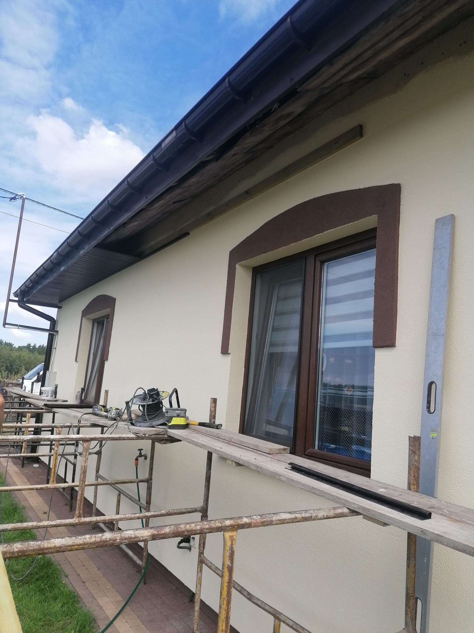 Elewacja domu z beżowym tynkiem, brązowymi obramowaniami okien i rusztowaniem, na którym leżą narzędzia i poziomica.