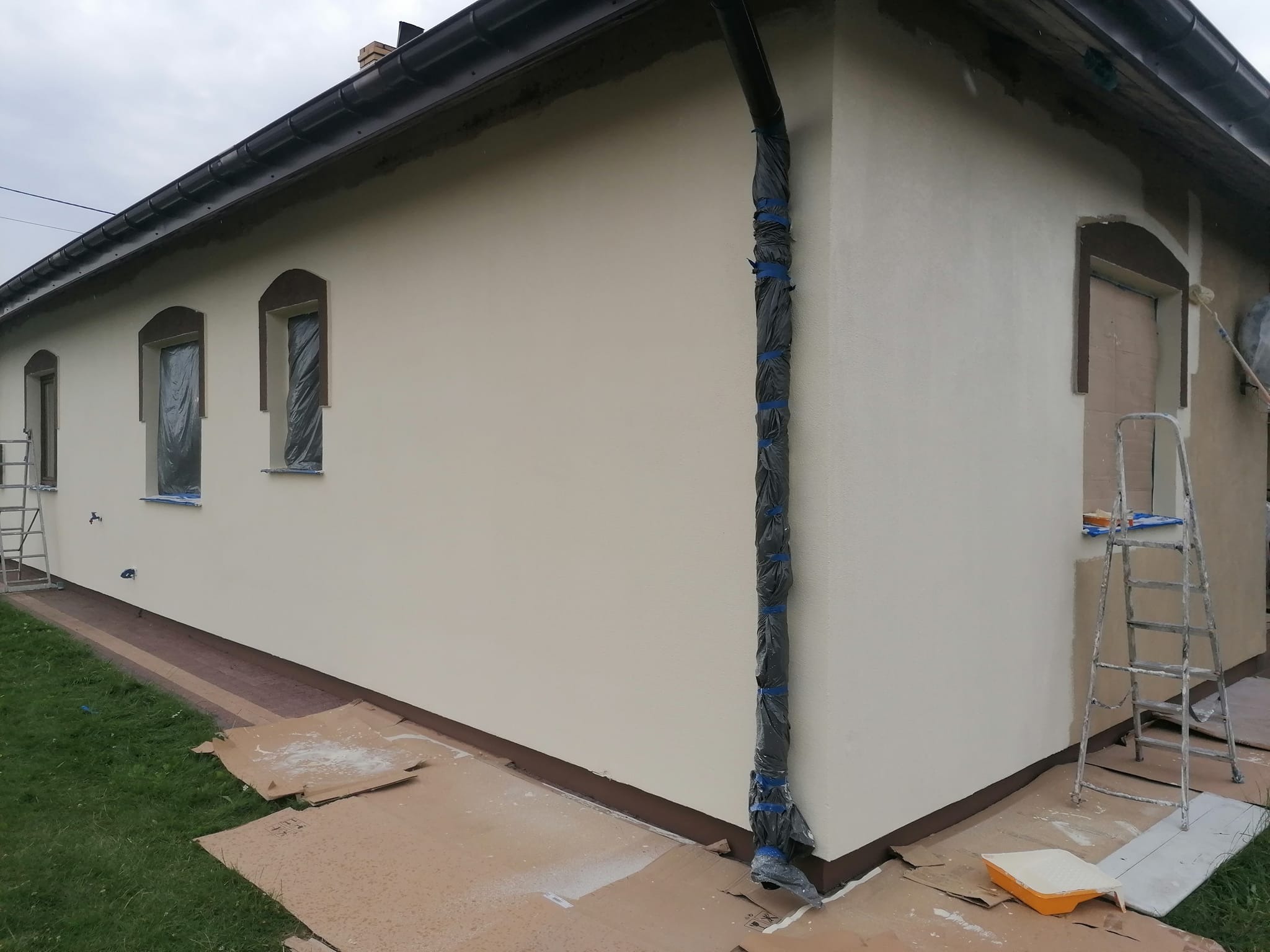 Elewacja domu jednorodzinnego w trakcie malowania, okna zabezpieczone folią, rynna owinięta czarną folią i taśmą, widoczne kartony ochronne na trawie, drabina oparta o ścianę.