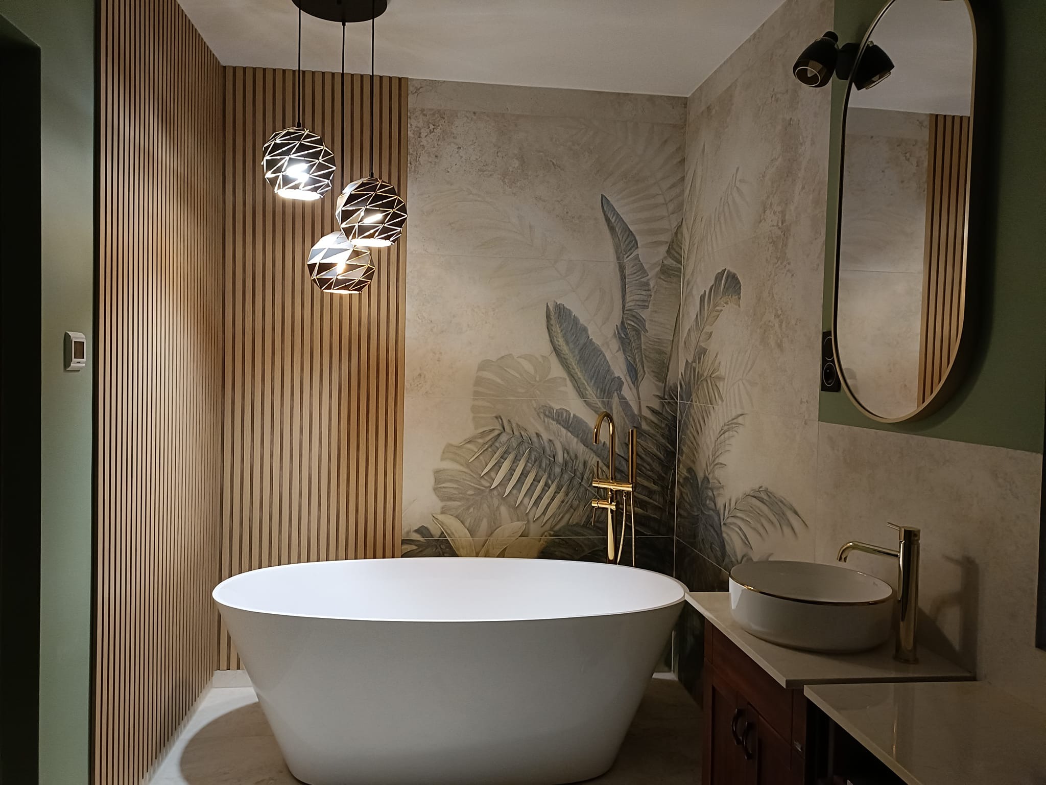 Nowoczesna łazienka z wolnostojącą wanną, armaturą w kolorze złota, płytkami z motywem roślinnym i drewnianymi lamelami na ścianie, oświetlona trzema designerskimi lampami wiszącymi.